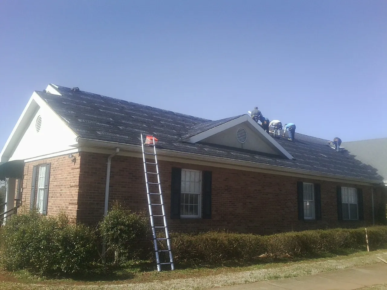 Crew tearing shingles off a brick ranch roof with a ladder set — Emergency Roof Repair work by our crew in Evans