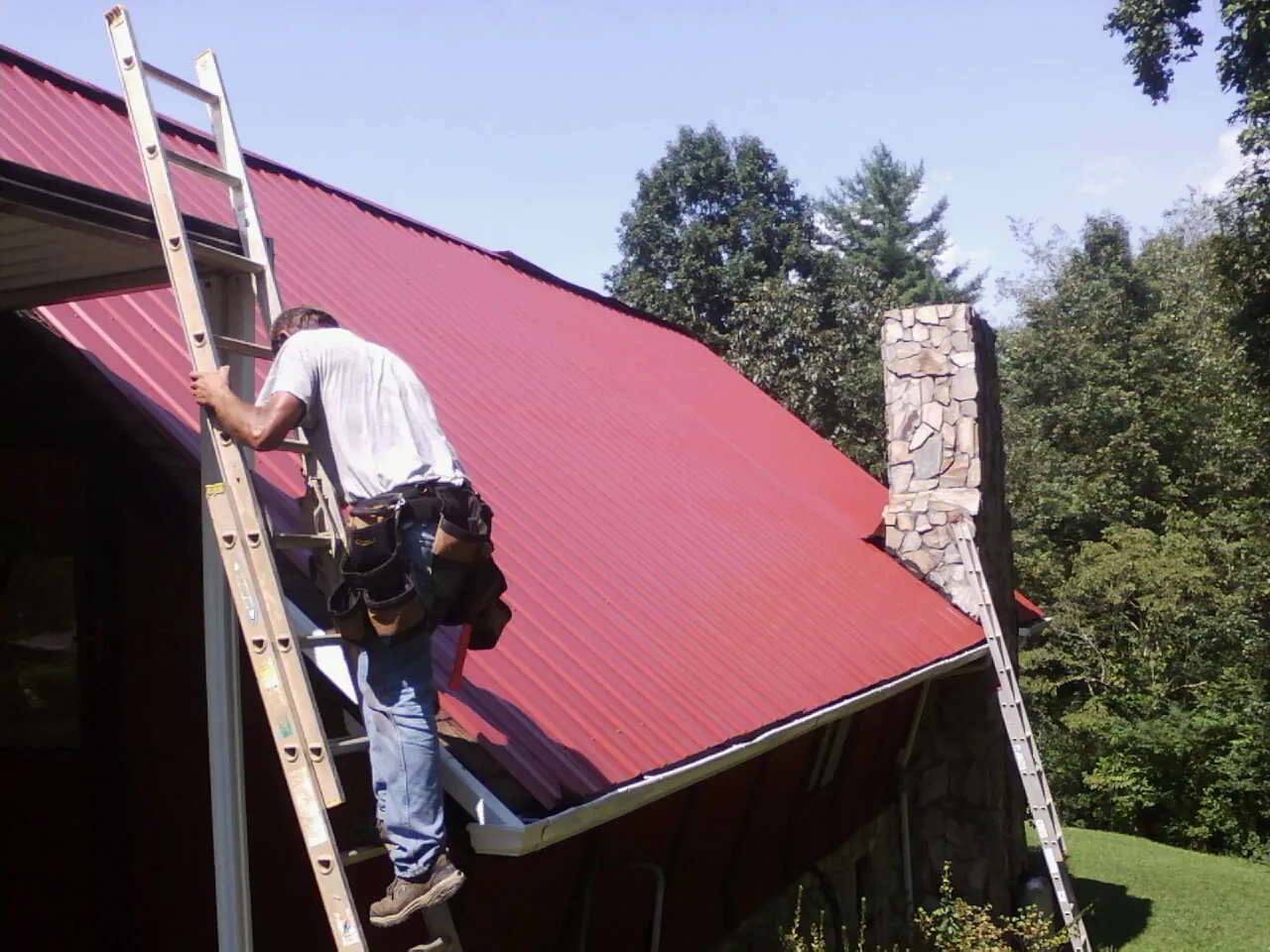 Roofer on a ladder beside a red metal roof and stone chimney — Soffit & Fascia Repair by Roofing Brothers in Evans